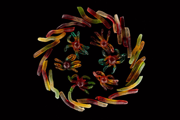 top view of colorful gummy spiders in circle of worms isolated on black, Halloween treat - 写真・画像