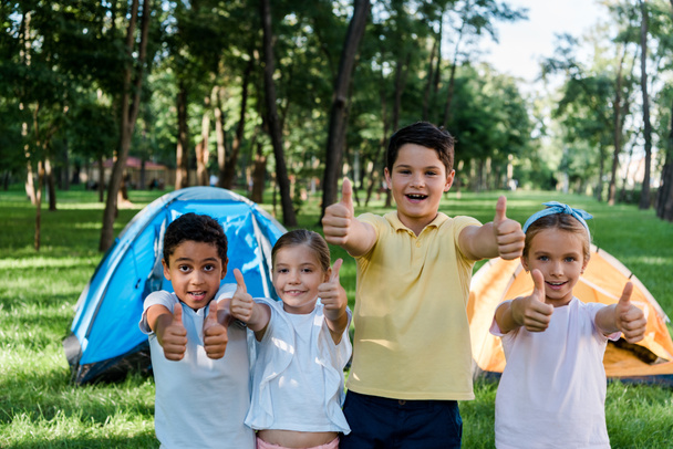 happy multicultural kids showing thumbs up near camps  - Fotó, kép
