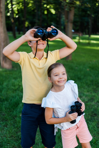 happy boy looking through binoculars near cheerful friend in park  - Фото, зображення