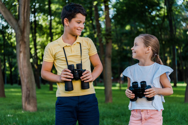 cheerful boy looking at cute friend holding binoculars in park  - Photo, Image