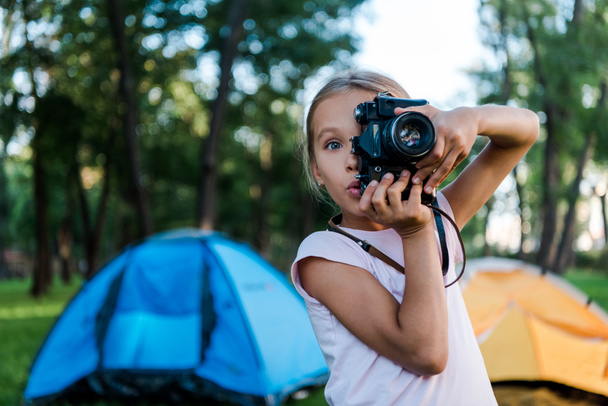 zaskoczony dzieciak trzyma aparat cyfrowy podczas robienia zdjęć w pobliżu obozów  - Zdjęcie, obraz