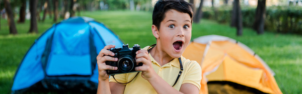 panoramic shot of surprised boy holding digital camera near camps in park  - Photo, Image