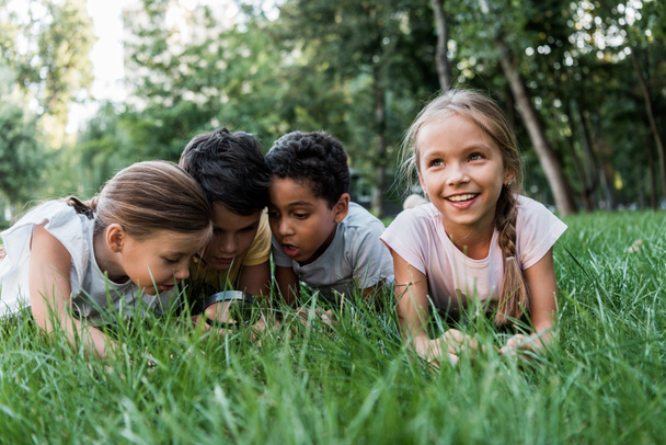 selective focus of cute multicultural children looking at green grass though magnifier  - Foto, imagen