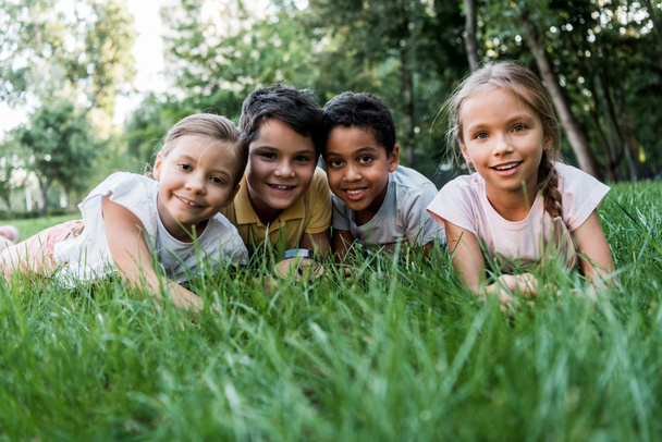 foco seletivo de crianças multiculturais bonitos sorrindo enquanto deitado na grama
  - Foto, Imagem