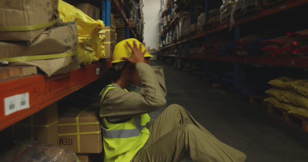 Side view close up of a young mixed race female warehouse worker taking off her hard hat and sitting down to take a break between storage shelves in a warehouse - Filmagem, Vídeo