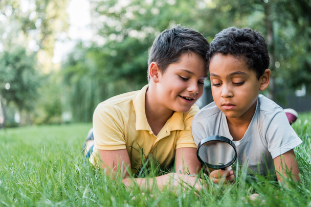 selective focus of adorable multicultural boys lying on green grass and looking through magnifier   - Photo, Image