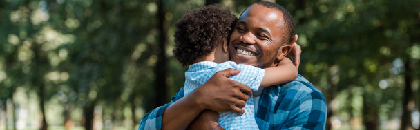 幸せなアフリカ系アメリカ人の父親が息子を抱きしめるパノラマショット  - 写真・画像