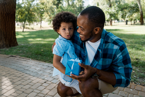 happy african american man holding blue gift and looking at curly son  - 写真・画像