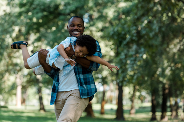 glimlachend Afro-Amerikaanse vader holding n armen schattige zoon in groen park  - Foto, afbeelding