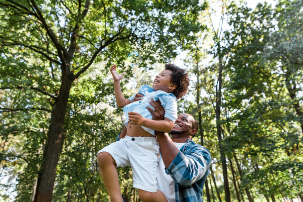 niski kąt widzenia szczęśliwego afrykańskiego amerykańskiego mężczyzny trzymania w ramionach cute syna w parku  - Zdjęcie, obraz
