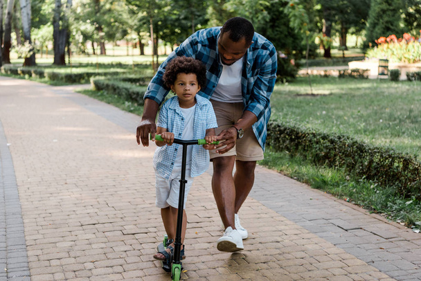bärtiger afrikanisch-amerikanischer Vater schaut Lockensohn beim Rollerfahren an  - Foto, Bild