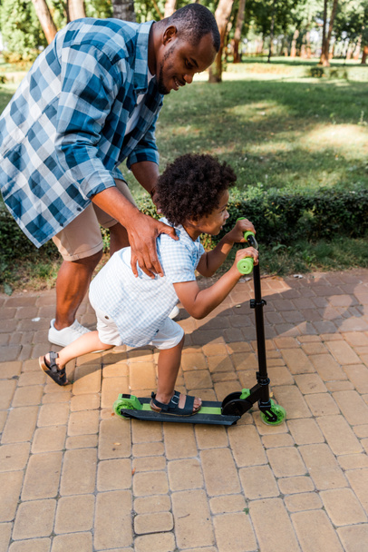 vista lateral do pai americano africano perto de criança feliz montando scooter
  - Foto, Imagem