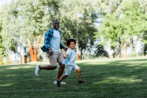 ευτυχισμένος Αφρικανός Αμερικανός πατέρας και γιος τρέχει σε γρασίδι - Φωτογραφία, εικόνα
