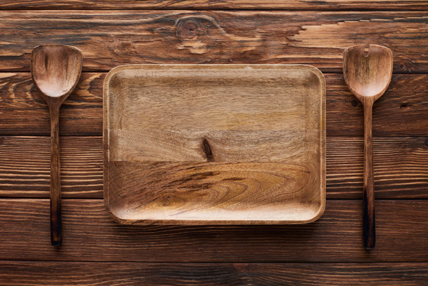 top view of empty rectangular dish between spoons on brown wooden surface with copy space - Foto, Imagem