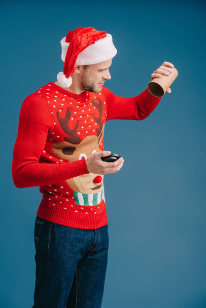 man in santa hat and christmas sweater holding empty cup of coffee, isolated on blue - Φωτογραφία, εικόνα