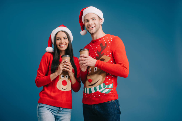 uśmiechnięta para w kapelusze Santa i Christmas Swetry trzymając kawę, aby przejść izolowane na niebiesko - Zdjęcie, obraz