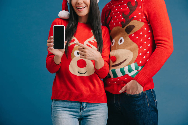 cropped view of couple in santa hats and christmas sweaters showing smartphone with blank screen isolated on blue - Photo, Image