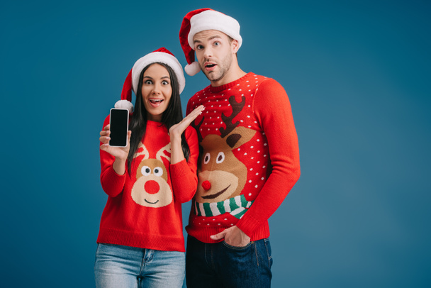 surprised couple in santa hats and christmas sweaters showing smartphone with blank screen isolated on blue - Фото, зображення