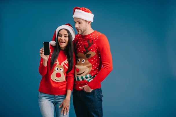 happy couple in santa hats and christmas sweaters showing smartphone with blank screen isolated on blue - Foto, afbeelding