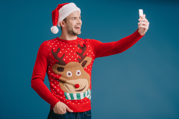 Красивый счастливый мужчина в шляпе Санты и рождественском свитере делает селфи на смартфоне, изолированный на голубом
 - Фото, изображение