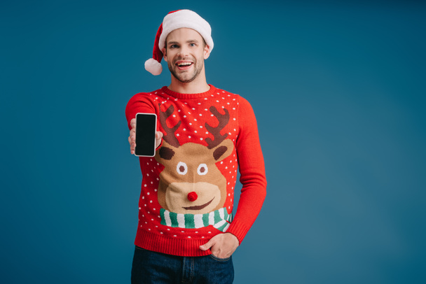 όμορφος άνθρωπος σε Σάντα καπέλο και χριστουγεννιάτικο πουλόβερ που δείχνει smartphone με κενή οθόνη, απομονωμένη στο μπλε - Φωτογραφία, εικόνα