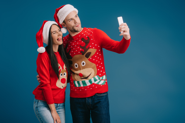 couple in santa hats and christmas sweaters sticking tongues out while taking selfie on smartphone isolated on blue - Photo, Image