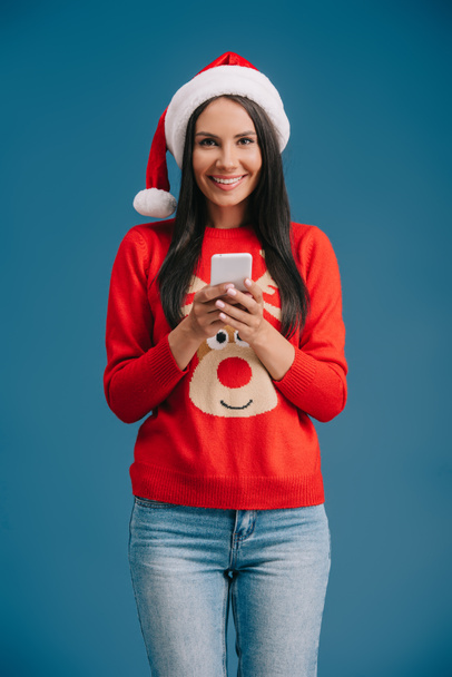 femme souriante en chapeau de Père Noël et pull de Noël en utilisant smartphone isolé sur bleu
 - Photo, image