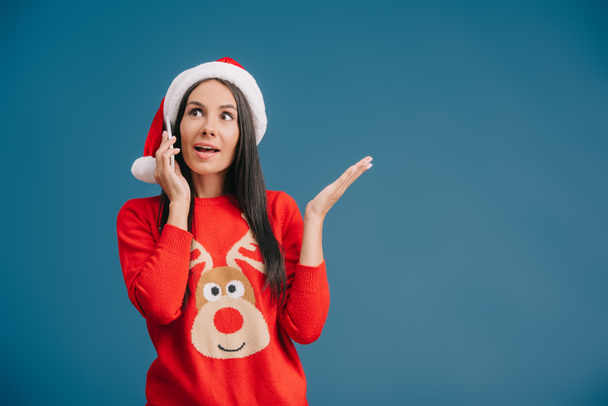 schöne Frau mit Weihnachtsmannmütze, die auf ihrem Smartphone isoliert auf blauem Grund spricht - Foto, Bild