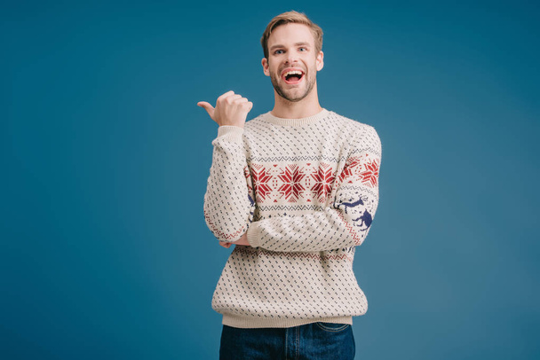 laughing man in winter sweater pointing at something isolated on blue - Fotoğraf, Görsel