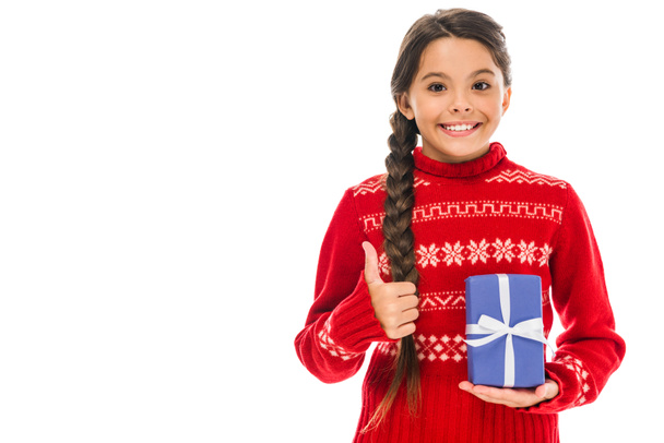 χαρούμενο παιδί με πουλόβερ που κρατάει το παρόν και δείχνει τον αντίχειρα του απομονωμένο στα λευκά  - Φωτογραφία, εικόνα