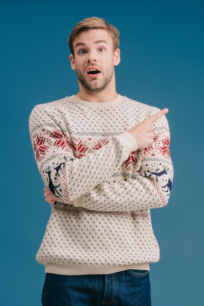 handsome shocked man in winter sweater pointing at something isolated on blue - Foto, imagen