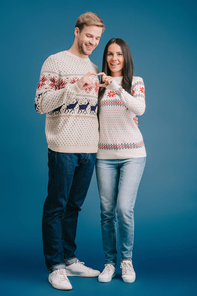 χαμογελαστά ζευγάρι σε χειμωνιάτικα πουλόβερ που δείχνουν το σημάδι της καρδιάς απομονωμένη στο μπλε - Φωτογραφία, εικόνα