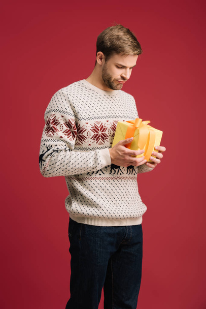 handsome thoughtful man holding christmas gift box isolated on red - Foto, immagini