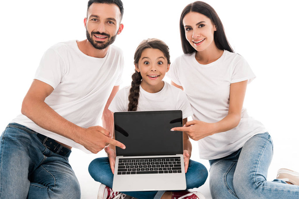 boldog szülők fehér pólóban ujjal mutogatnak a laptopra üres képernyővel a lánya közelében elszigetelt fehér  - Fotó, kép