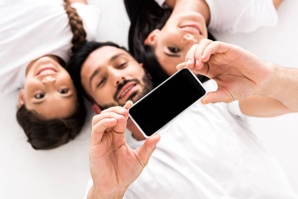 Draufsicht von glücklichen bärtigen Mann hält Smartphone mit leerem Bildschirm in der Nähe Frau und niedliches Kind isoliert auf weiß  - Foto, Bild