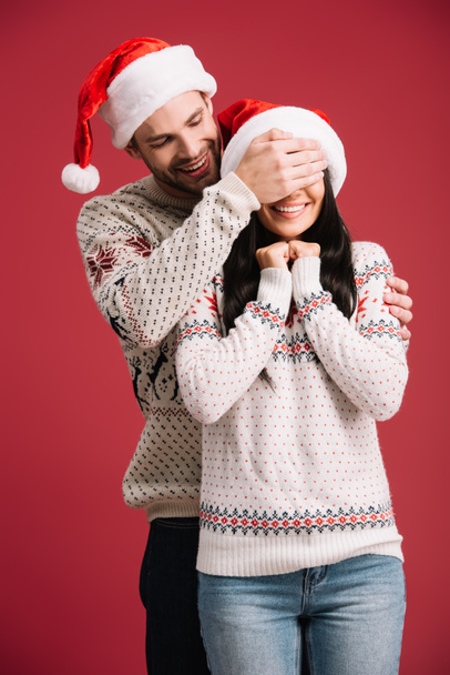 smiling man in santa hat closing eyes of happy girlfriend to make surprise, isolated on red - Фото, изображение