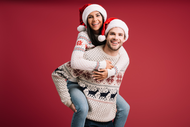 beautiful couple in santa hats and sweaters piggybacking isolated on red - Photo, Image