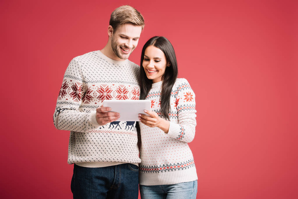 smiling couple in winter sweaters using digital tablet isolated on red - 写真・画像