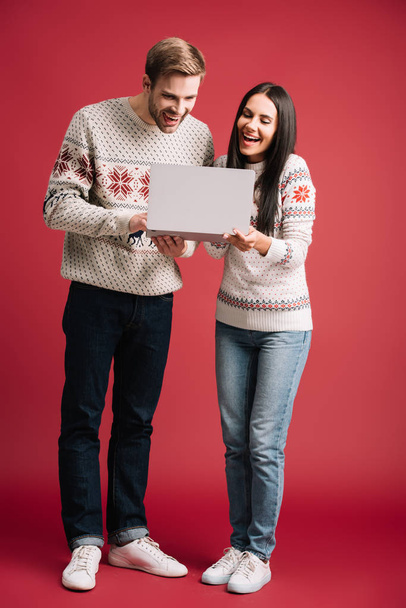 beautiful laughing couple in winter sweaters using laptop isolated on red - Fotografie, Obrázek