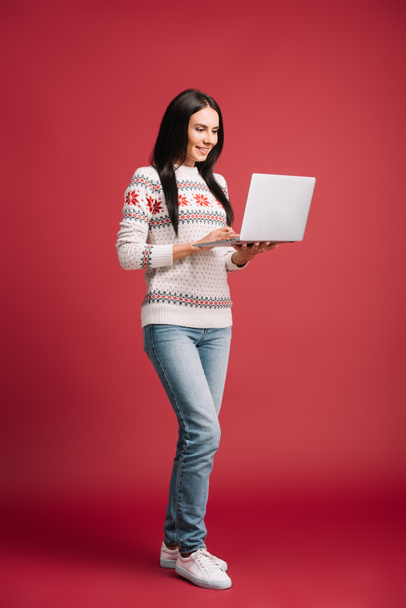 赤で隔離されたラップトップを使用して冬のセーターで笑顔の女性 - 写真・画像