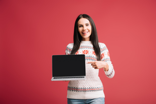 hermosa mujer en suéter de invierno apuntando a la computadora portátil con pantalla en blanco aislado en rojo
 - Foto, Imagen