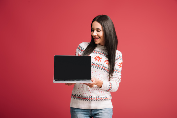smiling girl in winter sweater showing laptop with blank screen isolated on red - Φωτογραφία, εικόνα
