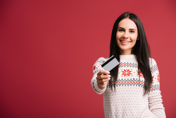 cheerful woman in winter sweater holding credit card, isolated on red  - Photo, Image