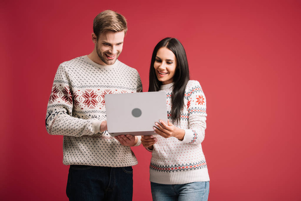 beautiful smiling couple in winter sweaters using laptop isolated on red - Photo, image