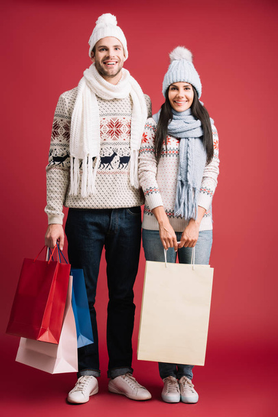 couple in winter sweaters, scarves and hats holding shopping bags isolated on red - Photo, Image