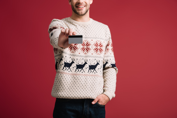bijgesneden weergave van de mens in winter trui met creditcard, geïsoleerd op rood  - Foto, afbeelding