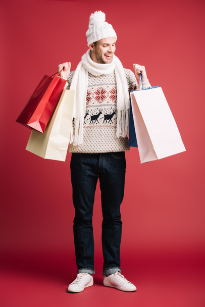cheerful man in winter scarf, hat and sweater holding shopping bags, isolated on red  - Fotoğraf, Görsel