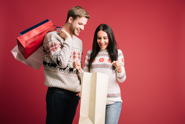 smiling couple in winter sweaters looking into shopping bags isolated on red - Valokuva, kuva