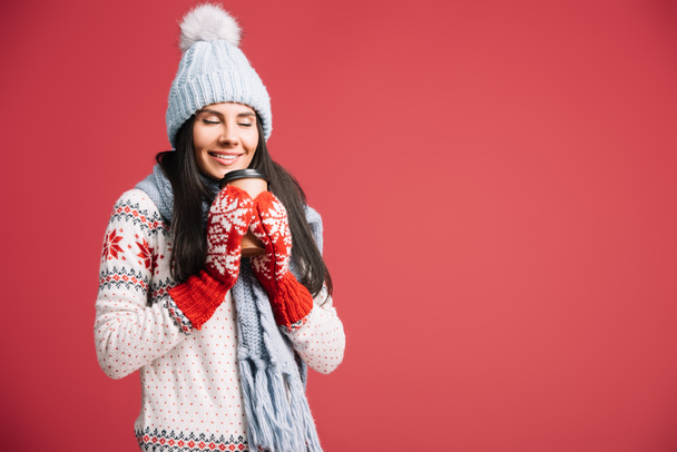 glückliche Frau mit Wintermütze, Schal und Handschuhen, die Coffee to go in der Hand halten, isoliert auf rot - Foto, Bild