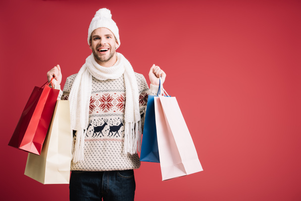 handsome smiling man in winter clothes holding shopping bags, isolated on red  - Fotó, kép